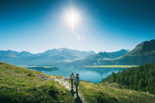 Hikers walking along a narrow trail overlooking the blue waters of Lake Sils